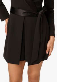 Robe noire portefeuille avec une ceinture en satin, des plis structurés, des manches longues et une texture lisse et mate. Mains partiellement visibles.