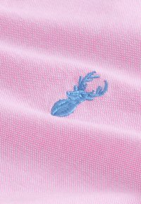 Tessuto rosa chiaro con un motivo testurizzato, caratterizzato da un emblema di cervo ricamato in blu. Il design è liscio e dettagliato.