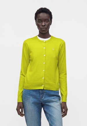 Femme portant un cardigan vert citron boutonné sur un haut blanc et un jean bleu, debout devant un fond blanc uni.