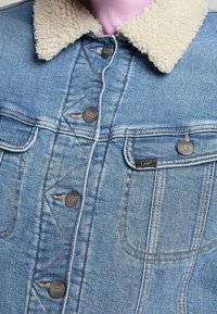 Džínová bunda světle modré barvy, s límcem z ovčí vlny, stříbrnými knoflíky a dvěma předními kapsami. Obsahuje kontrastní švy.