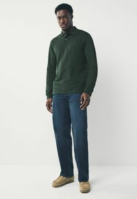 Dunkelgrünes Langarm-Polohemd, blaue Jeans und beige Schuhe. Das Polohemd hat einen Kragen und ein kleines Emblem auf der Brust.