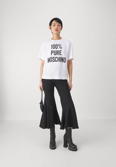 Balta trumpa rankovėmis marškinėliai su užrašu "100% PURŲ MOSCHINO" paryškintu juodu tekstu, derinami su juodomis platėjančiomis kelnėmis ir juodomis aukštakulnėmis aulinukėmis.