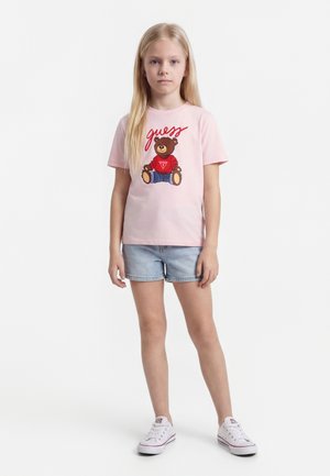 Νεαρή κοπέλα στέκεται στραμμένη προς τα εμπρός φορώντας ροζ t-shirt με σχέδιο αρκουδάκι, ανοιχτόχρωμα τζιν σορτς και λευκά αθλητικά παπούτσια σε λευκό φόντο.