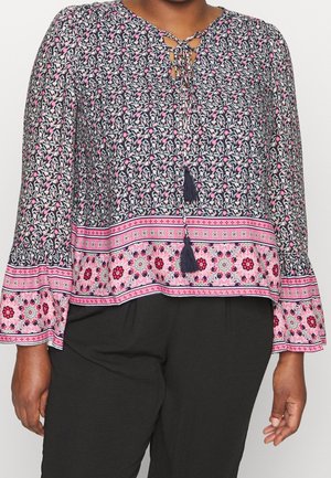 Mujer con blusa de estampado floral en tonos rosa, azul marino y blanco, con lazos con borlas en el escote y pantalones negros.