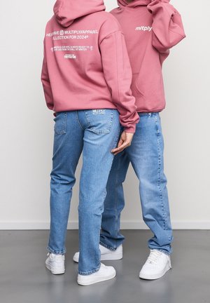 Twee mensen dragen roze hoodies en lichtblauwe jeans, staan dicht bij elkaar op een grijze vloer tegen een witte muur.