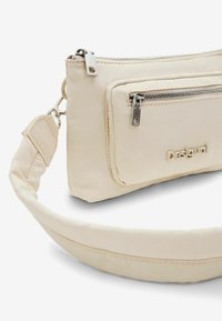 Beige crossbody bag made of soft material with a zipper pocket, smooth texture, and a padded strap. Features embossed logo on the front.