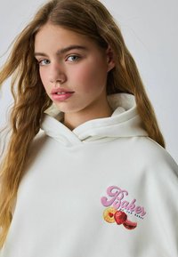 Valge kapuutsiga särk, mille ees on logo "Baker" roosade ja värvikate puuviljade graafikaga. Pehme tekstuur, reguleeritav kapuuts ja lõdva istuvusega.