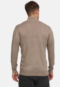 Pull gris clair avec un col montant, texture côtelée, manches longues et détails fendus sur les côtés, porté avec un pantalon foncé.