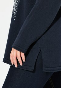 Navy gestrickter Pullover mit strukturiertem Stoff, seitlichen Schlitzern und verzierten Details auf der Vorderseite. Eine Hand, die am Saum ruht, zeigt die Passform.