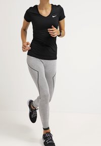 Czarna koszulka z krótkim rękawem z białym logo, w zestawie z szarymi legginsami z czarnymi akcentami. Buty sportowe są czarne z białymi detalami.