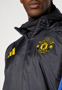 Schwarze wasserabweisende Jacke mit gelbem Manchester United-Logo, Reißverschluss vorne und dezenter Textur sowie hohem Kragen-Design.