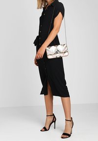 Vestido negro abotonado con mangas cortas, cintura ajustada y una abertura. Incluye un bolso de mano metálico con patrón y tacones altos negros con tiras.