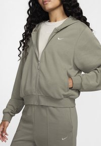 Grønn hettejakke med glidelås og avslappet passform, laget av mykt stoff. Har hette, frontlommer og liten hvit Nike-logo på brystet.
