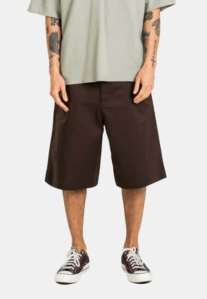 Man wearing brown knee-length shorts, light gray oversized t-shirt, and dark brown low-top sneakers with white laces and soles.