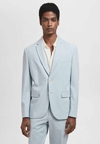 Jasnoniebieski garnitur wykonany z miękkiego materiału, z wyłamią klapą, dwiema przednimi kieszeniami i jasnymi guzikami. Noszony z kremową koszulą.