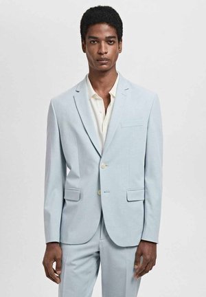 Abito blu chiaro in tessuto morbido, con revers a punta, due tasche frontali e bottoni chiari. Indossato con una camicia color crema.
