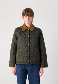 Gequilted groene jas met een bruine suède kraag, gouden knopen en bruine zakaccenten. Het model draagt een spijkerbroek en staat voor een neutrale achtergrond.