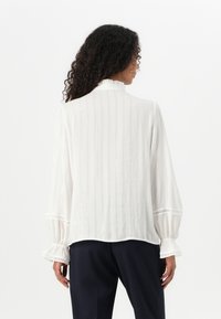 Vit blus med en texturerad tyg med vertikala ränder. Puffärm med elastiska manchetter och dekorativ spetskant. Slät yta.