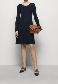 Námořnické úpletové šaty s dlouhými rukávy, texturovaným vzorem a vlnitým lemem, doplněné hnědou kabelkou přes rameno a černými lodičkami na podpatku s přezkami.