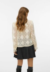 Koronkowa bluzka z misternym wzorem w diamenty w odcieniu jasnej bieli, posiadająca długie rękawy i dekoracyjne mankiety, zestawiona z ciemnoszarym spódnicą.