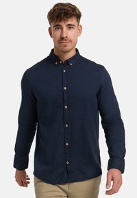 Mann mit kurzen lockigen Haaren, der ein marineblaues Hemd mit Knöpfen und beige Hosen trägt, vor einem weißen Hintergrund stehend.