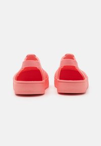 Sandálias cor-de-rosa coral com parte traseira aberta, tira elástica, parte superior texturizada e sola de borracha sólida, apresentando um acabamento suave e um design simples.