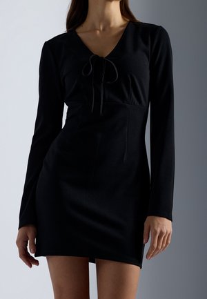 Woman wearing a fitted black long-sleeve mini dress with a V-neck and tie detail at the chest, standing against a light gray background.