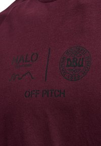 Vinröd t-shirt med svart tryckt text. Visar "HALO" och "OFF PITCH" med en cirkulär logotyp. Mjuk bomullsmaterial med rundad hals.