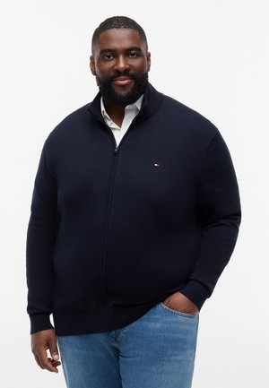 Muž s bradou v tmavomodrom svetri na zips, oblečený cez bielu košeľu a modré džínsy, stojaci s jednou rukou v vrecku na bielom pozadí.