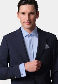 Navy suit jacket over light blue dress shirt with button detail. Pocket square features a geometric pattern. Cufflinks visible on the shirt sleeves.