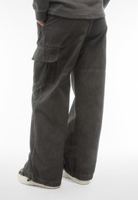 Pantaloni cargo in tessuto grigio scuro, vestibilità ampia con tasche laterali. Presentano dettagli di cucitura e polsini elasticizzati. Indossati con sneakers di colore chiaro.