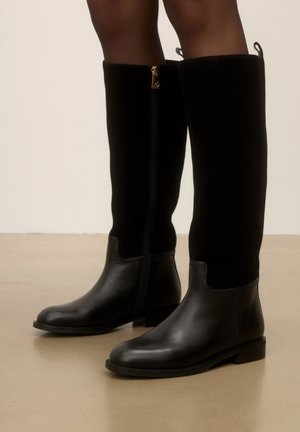 Bottes hautes noires avec partie inférieure en cuir et partie supérieure en daim, fermeture éclair sur le côté intérieur, portées sur jambes nues sur fond neutre.