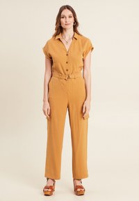 Mosterdgele jumpsuit met een kraag, knoopsluiting aan de bovenkant, ceintuur in de taille, met brede pijpen en zijzakken. Houten plateau schoenen.