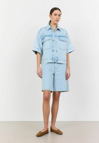 Svetlo modra denim srajca z oversize rokavi in dvema žepoma na prsih, v kombinaciji z usklajenimi kratkimi hlačami, oboje z jasno, minimalistično zasnovo.