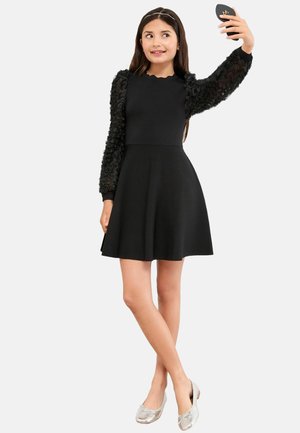 Abito nero con un corpetto aderente e una gonna svasata, caratterizzato da maniche floreali nere testurizzate. Le ballerine argento completano l'outfit.