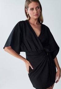 Crna haljina na preklop s širokim kimono rukavima, dubokim V-izrezom, nabranim pojasom s metalnim detaljima i glatkom teksturom tkanine.