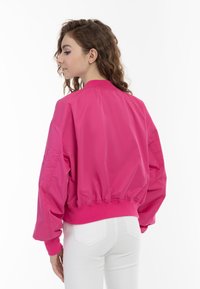Giacca bomber fucsia brillante con fascia in vita e polsini a coste, tessuto lucido e vestibilità oversize. Presenta spalle scese e tasche laterali.