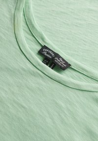 Jasnozielona koszulka z miękkiego materiału z okrągłym dekoltem. Zawiera metkę z informacjami o marce "Superdry" oraz rozmiarze.