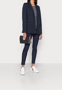 Blazer azul marino, camiseta de rayas, jeans oscuros, zapatillas blancas. Bolso negro con herraje dorado. Conjunto casual con diseño moderno y líneas limpias.