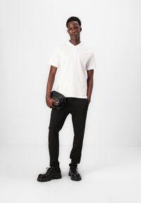 Maglietta polo bianca, pantaloni a righe nere e scarpe nere spesse. Tiene una piccola borsa nera. Design minimalista su uno sfondo semplice.