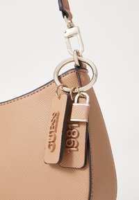 Bolso texturizado beige con herrajes dorados, que presenta detalles de llavero y etiquetas grabadas con "GUESS" y "1981". Forma redondeada.