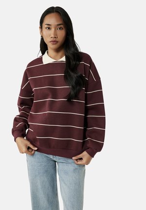 Ung kvinde med langt mørkt hår iført en vinrød sweatshirt med striber over en hvid skjorte med krave og lyseblå jeans, står med hænderne nær livet.