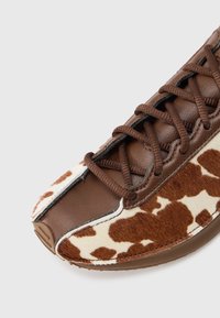 Pruun nahast king, millel on lehmanahast küljepaneel. Omab pruune paelu ja tekstureeritud materjali, rõhutades ainulaadset disaini ja kuju.