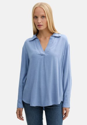 Femme blonde portant une blouse ample à manches longues bleu clair avec un petit col et un jean bleu foncé sur un fond uni.