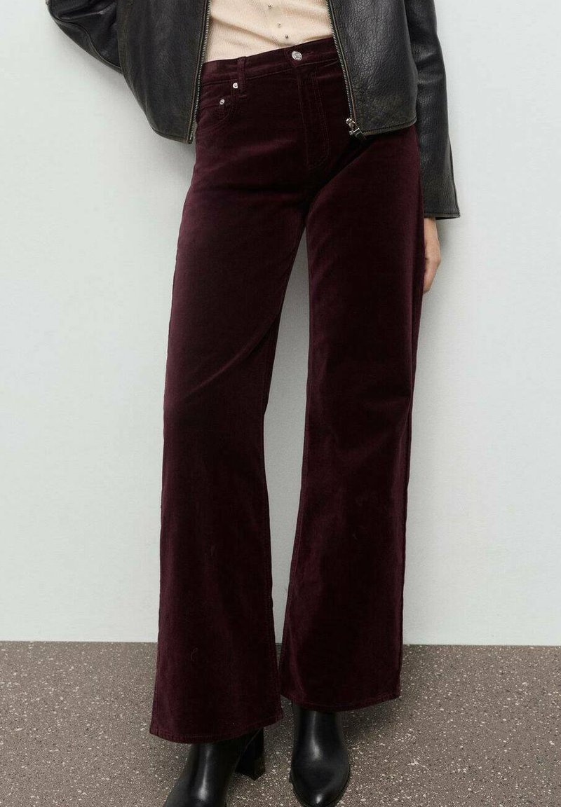 Pantalones de pana burdeos de pierna ancha con textura suave, bolsillos frontales y detalles de metal plateado; combinados con botas negras hasta el tobillo y una chaqueta de cuero.