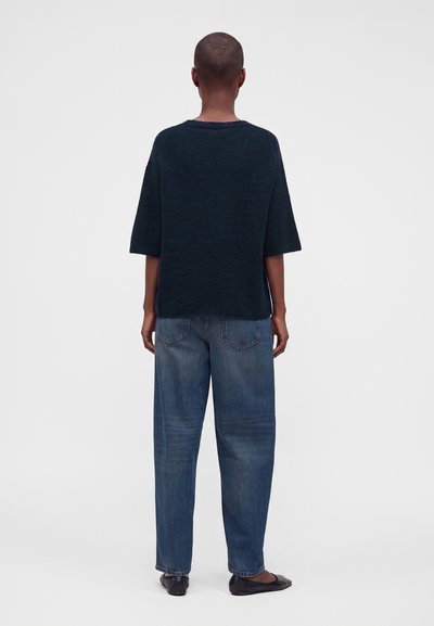 Personne aux cheveux courts portant un haut ample sombre à manches courtes, un jean bleu et des ballerines noires, debout de dos sur un fond uni.