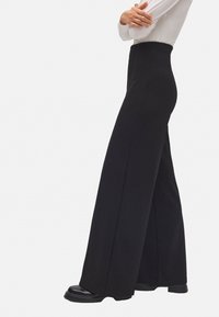 Mujer de pie con los brazos cruzados, vistiendo pantalones anchos de tiro alto negros, una blusa blanca de manga larga y zapatos negros, sobre un fondo blanco liso.