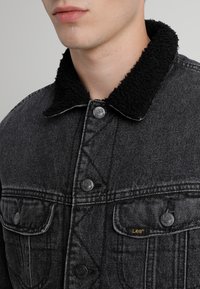 Black denim jacket with a fluffy black collar, button closure, and front pockets. Features subtle brand tag in gold on the chest pocket.