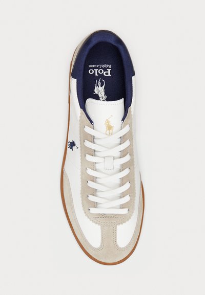 Zapatilla de cuero blanca con acentos de ante beige, interior azul marino, cordones blancos y detalles de logotipo dorado y azul marino en los laterales.