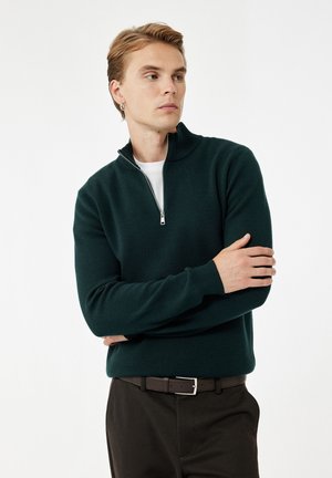 Pull à col zippé vert foncé avec des manches longues, des poignets côtelés et une texture lisse. Porté sur un t-shirt blanc et associé à un pantalon sombre.